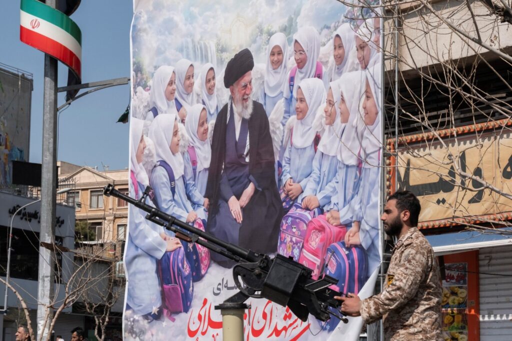 Ein iranischer Revolutionsgardist vor einem Poster mit dem bei einem israelischen Enthauptungsschlag getöteten geistlichen Führer Ayatollah Ali Chamenei und Schulmädchen, die angeblich ebenfalls bei einem Luftangriff ums Leben kamen. 