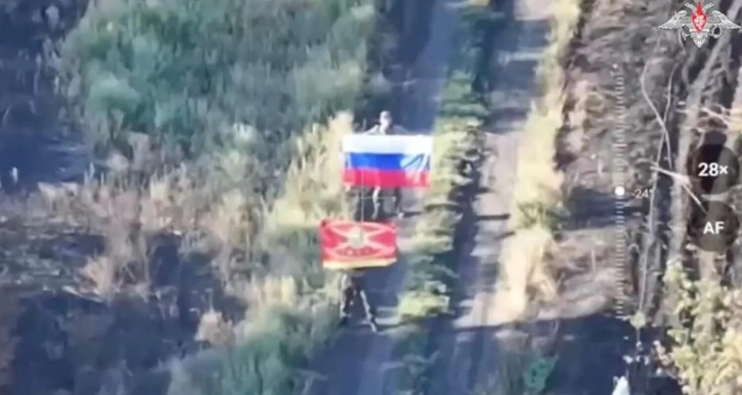 Lächeln für die Drohne: Zwei russische Soldaten posieren mit Flaggen für ein Beweisfoto von der Eroberung eines Dorfes in der Ukraine.